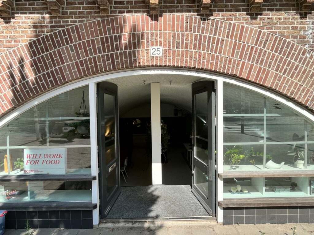 Ingang van een gebogen bakstenen pand met glazen etalages en het huisnummer 25, getiteld "Tussen de Bogen".